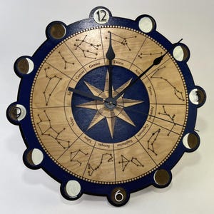 May include: A wooden wall clock with a navy blue border and a light brown face. The clock features a compass rose design at the center, surrounded by zodiac constellations and the names of the zodiac signs. The clock hands are black.