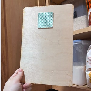 May include: A light-colored wooden board with a square of green and white adhesive tape attached. The board is rectangular with rounded corners and appears to be held open against a wooden cabinet. The background includes shelves with food storage containers.
