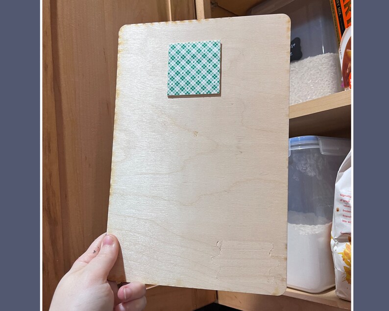 May include: A light-colored wooden board with a square of green and white adhesive tape attached. The board is rectangular with rounded corners and appears to be held open against a wooden cabinet. The background includes shelves with food storage containers.