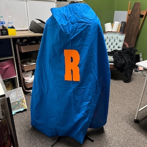 May include: A blue cape with an orange letter "R" on the back. The cape is hanging on a stand.