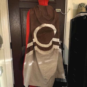 Könnte beinhalten: Ein Kapuzenmantel mit braunem und weißem Design, getragen über einem roten Outfit. Der Umhang hat einen großen weißen Kreis mit braunem Umriss und horizontalen Streifen. Das Outfit umfasst eine rote Kapuze, ein langärmliges Hemd und Leggings.