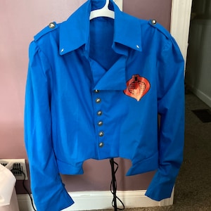 Puede incluir: Una camisa azul de manga larga con un emblema de Cobra en la parte delantera. La camisa tiene un cuello alto y un dobladillo recortado.