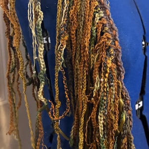 Puede incluir: Primer plano de una bufanda texturizada hecha a mano en tonos marrón, naranja, verde y amarillo. La bufanda parece estar hecha de hilo y tiene un diseño artístico único. La bufanda está drapeada sobre un maniquí azul.