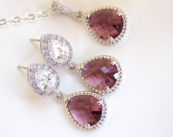 Puede incluir: Un conjunto de pendientes y un collar con cristales en forma de lágrima. Los pendientes tienen un cristal transparente más pequeño rodeado por un cristal morado más grande. El collar tiene un solo cristal morado más grande.