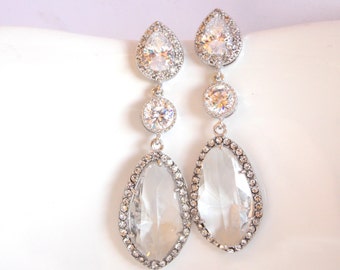 May include: A pair of silver teardrop earrings with clear crystal stones. The earrings have a large teardrop-shaped stone surrounded by smaller stones, and a smaller round stone above it.