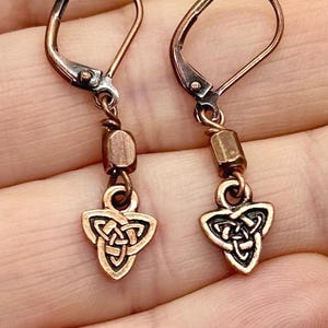 Könnte beinhalten: Ein Paar kupferfarbene Ohrringe mit keltischem Knoten. Jeder Ohrring hat einen Klappverschluss, eine kleine rechteckige Perle und einen dreieckigen Knotenanhänger. Die Ohrringe sind mit einem Antik-Finish versehen.