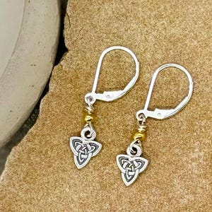 May include: A pair of silver-toned leverback earrings with a Celtic knot design. The earrings have a small gold-colored bead accent. The earrings are displayed on a textured, tan surface.