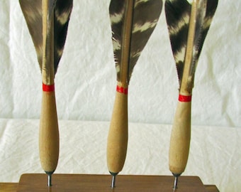 May include: Three vintage darts with wooden handles and feathered flights. Each dart has a red band near the flight and a metal tip. The darts are displayed in a dark wooden holder, showcasing their classic design and craftsmanship.