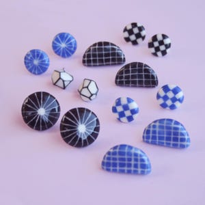 Può includere: Un set di 10 orecchini in ceramica con varie forme e motivi geometrici. Gli orecchini sono in tonalità di blu e nero con accenti bianchi. Gli orecchini sono esposti su uno sfondo viola chiaro.