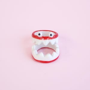 Peut inclure: Une bague en céramique blanche avec une bordure rouge et une grande bouche ouverte avec des dents blanches. La bague a deux yeux noirs.