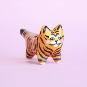 以下が含まれることがあります： 黒とオレンジのストライプのセラミック製の虎の置物。虎は、ピンクの頬と黒い目をした、かわいい漫画のような顔をしています。