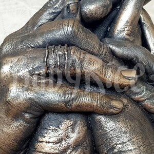 May include: A bronze sculpture of two hands clasped together, with a ring on one of the fingers. The sculpture is detailed with textured skin and fingernails.