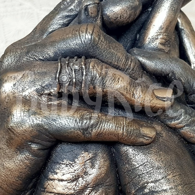 May include: A bronze sculpture of two hands clasped together, with a ring on one of the fingers. The sculpture is detailed with textured skin and fingernails.