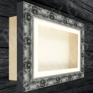 Könnte beinhalten: Ein silberner, verzierter Bilderrahmen mit einem weißen Passepartout und einer Holzrückseite. Der Rahmen hat ein dekoratives, erhabenes Design.