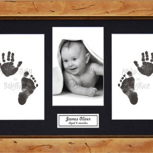 May include: A black and white photograph of a baby with a hand and footprint imprint. The baby is lying on a white blanket and looking at the camera. The photo is framed in a wooden frame with a black mount. The text "James Oliver Aged 6 months" is printed below the photo.