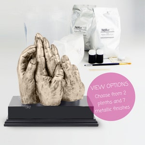 May include: A three-dimensional hand casting kit with three hands clasped together. The casting is on a black base. The kit includes two bags of white powder, two small bottles of black liquid, a small bottle of white liquid, a paintbrush, two wooden sticks, and a piece of white fabric. The text "VIEW OPTIONS Choose from 2 Plinths and 7 metallic finishes" is on a pink circle.