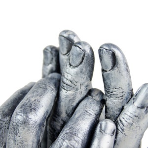 May include: A silver-colored handprint casting of two hands, likely a baby's and an adult's, with the fingers intertwined. The casting is detailed, showing the individual ridges and lines of the fingers.
