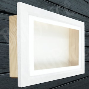 Könnte beinhalten: Weiße Holz-Schattenbox mit weißem Passepartout und einer transparenten Glasfront. Die Box ist auf einem dunklen Holzgrund montiert.