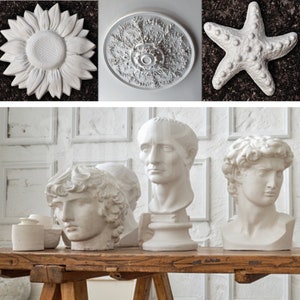 May include: Four white plaster sculptures: a sunflower, a decorative ceiling medallion, a starfish, and three busts of people. The busts are displayed on a wooden table.