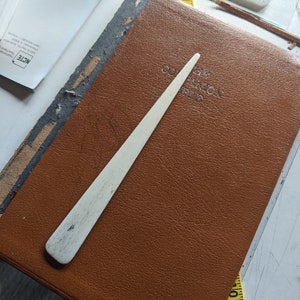 Könnte beinhalten: Nahaufnahme eines braunen, ledergebundenen Buches mit dem geprägten Text "The Caravan Club" auf dem Einband. Ein weißes, knochenartiges Papiermesser liegt auf dem Buch.