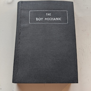 May include: A black hardcover book with the title "THE BOY MECHANIC" printed in white on the front cover.