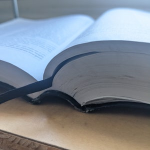 May include: An open book with white pages and a black bookmark. The book is resting on a light brown wooden surface. The pages are slightly yellowed, suggesting age or frequent use. The image is lit by natural light.