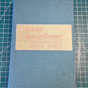 May include: The cover of a blue cookbook titled "The New Fannie Farmer Boston Cooking-School Cook Book."