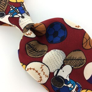 May include: A red silk tie with a pattern of various sports balls and a cartoon character. The balls include baseballs, basketballs, soccer balls, and golf balls. The cartoon character is wearing a blue shirt and holding a golf club.
