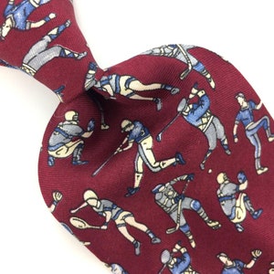 May include: A red silk tie with a repeating pattern of men playing different sports. The men are wearing blue and white uniforms and are depicted in various poses. The tie is a classic design and would be a great addition to any man's wardrobe.