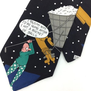 May include: A black tie with a cartoon illustration of a golfer hitting a bucket of golf balls. The text on the tie reads "I told my wife I'd be home after I hit a bucket of balls."