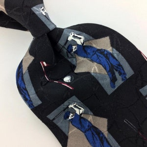 May include: A black silk tie with a repeating pattern of golfers in blue and white, holding golf clubs and golf balls. The background is a black and gray geometric pattern.