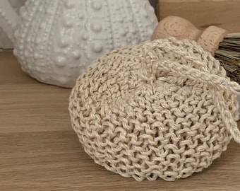 Op de afbeelding: Een ronde, beige, handgebreide katoenen afwasspons met een klein lusje om op te hangen. De spons heeft een gestructureerd patroon met open steken en is gemaakt van garen in een natuurlijke kleur voor het reinigen van oppervlakken.