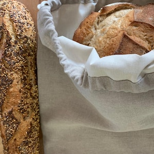Peut inclure: Un sac à pain en lin beige avec un cordon de serrage et une doublure intérieure blanche, contenant une miche de pain au levain croustillante. À côté du sac se trouve une baguette longue et fine recouverte de graines variées.