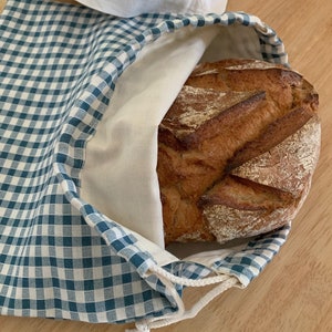 Peut inclure: Un sac à pain en tissu avec un motif à carreaux vichy bleu et blanc à l'extérieur et une doublure intérieure blanc cassé. Le sac est doté d'une fermeture à cordon avec des liens blancs et contient une grosse miche de pain au levain croustillante.