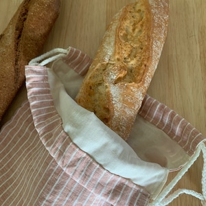 Könnte beinhalten: Zwei Baguettes, eines in einer wiederverwendbaren Brottasche. Die Tasche besteht aus einem rosa-weiß gestreiften Stoff mit Kordelzug. Das andere Baguette liegt auf einer Holzoberfläche.
