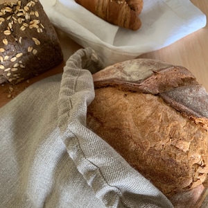 Op de afbeelding: Een rond brood met een bruine korst zit in een beige linnen tas met trekkoord. Daarnaast zijn een donkerbruin rechthoekig brood met havermout en twee goudbruine croissants op een witte doek op de achtergrond zichtbaar.