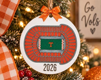May include: A round white wooden Christmas ornament featuring a graphic of a football stadium with orange seating and a green field with a central orange T. The year 2026 is printed in black at the bottom, and an orange ribbon bow is attached at the top.