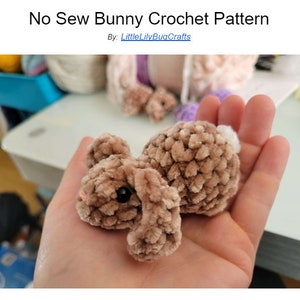 May include: A brown crocheted bunny with a white tail, black eyes, and a pink nose. The bunny is lying on its side in a person's hand.
