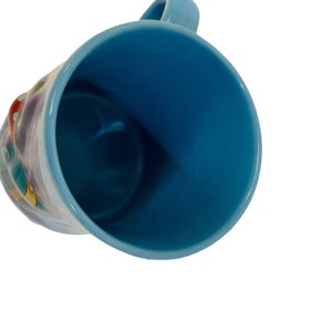 May include: A light blue ceramic mug with a matching handle. The interior of the mug is a darker shade of blue. The exterior features a colorful illustration. The mug is set against a white background.