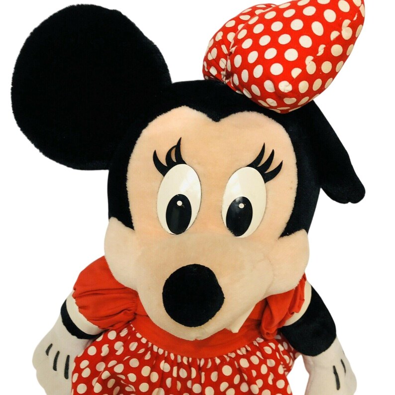 May include: A plush Minnie Mouse doll with black ears, a red and white polka dot bow, and a red polka dot dress. The doll has large eyes and a black nose. The doll's arms and hands are white.