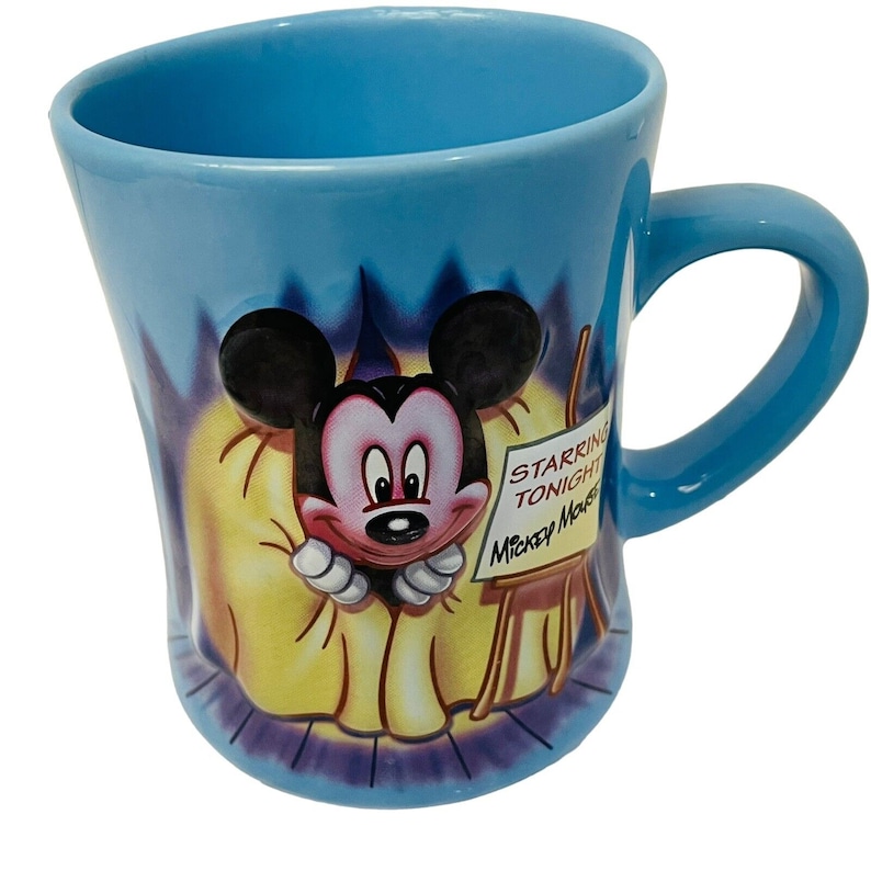 May include: A light blue ceramic mug featuring Mickey Mouse. The mug has a curved shape and a handle. Mickey Mouse is depicted in a 3D design, with a sign that reads "Starring Tonight Mickey Mouse".