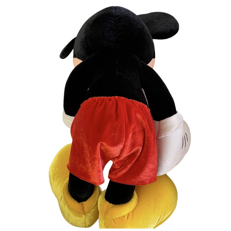 Puede incluir: Un peluche de Mickey Mouse, visto desde atr&aacute;s. El juguete presenta un cuerpo negro, pantalones cortos rojos, guantes blancos y zapatos amarillos. Las orejas son grandes y negras. El peluche es suave y tierno.