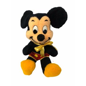 May include: A plush Mickey Mouse doll with black fur, a peach-colored face, and large black ears. The doll has a yellow bow, red shorts with yellow buttons, and yellow feet. A classic Disney character.