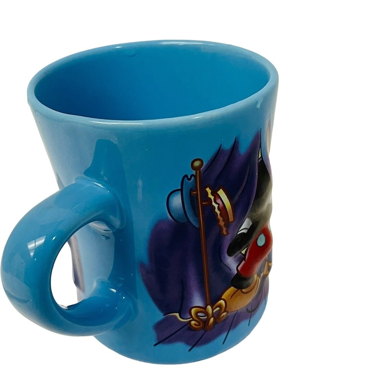 May include: A light blue ceramic mug with a curved handle. The mug features a cartoon character design with black, red, and gold accents. The design is set against a dark purple background.
