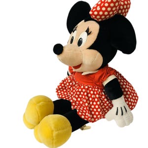May include: A plush Minnie Mouse doll, featuring black ears, a red and white polka dot bow, and a matching dress. The doll has a cream-colored face, black eyes, and yellow shoes. The doll is sitting down with its arms at its sides.