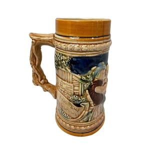 以下が含まれることがあります： 茶色のハンドルと蓋が付いた、ヴィンテージのセラミックビールジョッキ。本体には、人物、建物、葉が描かれた詳細なレリーフシーンがあり、バナーには「Froh auf leben will」という言葉が書かれています。ジョッキはタンと茶色の配色です。