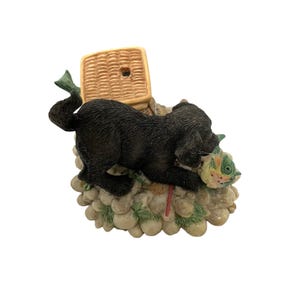 May include: A figurine of a black dog with a fish in its mouth, next to a woven basket. The dog is positioned on a rocky base with green and beige accents. The fish is multi-colored.