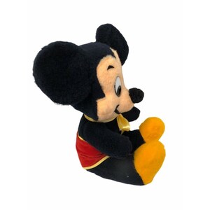 May include: A plush Mickey Mouse toy in a seated position. The toy has black fur, a tan face, and large black ears. Mickey wears a red vest with yellow trim, yellow gloves, and a yellow bow tie. The toy is set against a white background.