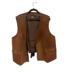 May include: A brown suede vest with a brown lining and a dark brown collar. The vest has two front pockets and is hanging on a wooden hanger. The vest is unbuttoned, revealing the interior.