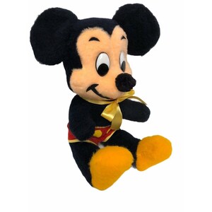 May include: A vintage Mickey Mouse plush toy. The plush features black fur, a tan face, and large black ears. Mickey wears red shorts with yellow buttons, yellow shoes, and a yellow bow tie. The toy is sitting with its legs extended.